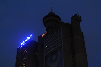 A dark, towering building with illuminated signage reading 'Zenter'. The structure has a rounded top with antennas and observation deck features. There is a visible image or artwork on the side depicting a simplistic human figure.