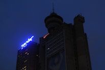 A dark, towering building with illuminated signage reading 'Zenter'. The structure has a rounded top with antennas and observation deck features. There is a visible image or artwork on the side depicting a simplistic human figure.
