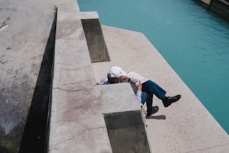 A person wearing a white hat and holding a newspaper is sitting on a concrete structure near a body of blue water. The sun casts shadows on the textured surface of the concrete, and the scene appears calm and relaxed.