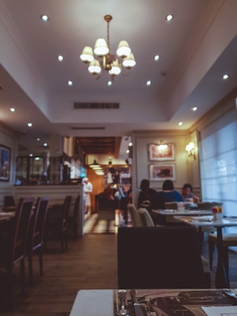 A cozy restaurant interior featuring a warm, ambient lighting with a central chandelier. The room has tables set for dining, some occupied by people. The setting is relaxed and inviting, with decor including framed pictures on the walls. The lighting creates a soft, intimate atmosphere.