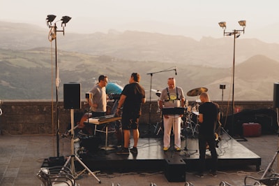 A group of five people gathered on an outdoor stage set up with musical instruments, such as keyboards and drums, with a scenic mountain landscape in the background during sunset. The individuals appear to be preparing for a performance, surrounded by lighting and sound equipment.