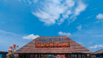 A thatched entrance sign with brightly colored lettering spelling 'KURIFTU WATER PARK' against a vivid blue sky with scattered clouds. In the background, part of the water park with slides and structures is visible.