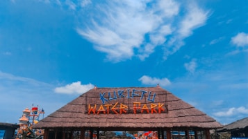 A thatched entrance sign with brightly colored lettering spelling 'KURIFTU WATER PARK' against a vivid blue sky with scattered clouds. In the background, part of the water park with slides and structures is visible.