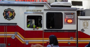 A red and white emergency vehicle with the FDNY HazMat logo and 'Specialists' written on its side. A person is visible inside the vehicle, and there is a pedestrian in the foreground.