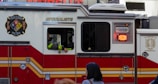 A red and white emergency vehicle with the FDNY HazMat logo and 'Specialists' written on its side. A person is visible inside the vehicle, and there is a pedestrian in the foreground.