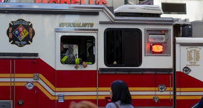 A red and white emergency vehicle with the FDNY HazMat logo and 'Specialists' written on its side. A person is visible inside the vehicle, and there is a pedestrian in the foreground.