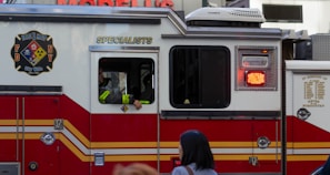 A red and white emergency vehicle with the FDNY HazMat logo and 'Specialists' written on its side. A person is visible inside the vehicle, and there is a pedestrian in the foreground.