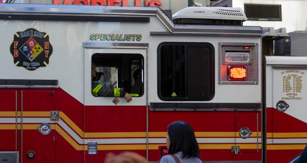 A red and white emergency vehicle with the FDNY HazMat logo and 'Specialists' written on its side. A person is visible inside the vehicle, and there is a pedestrian in the foreground.