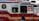 A red and white emergency vehicle with the FDNY HazMat logo and 'Specialists' written on its side. A person is visible inside the vehicle, and there is a pedestrian in the foreground.