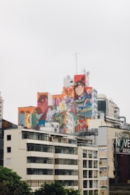 A cityscape featuring a building with a large, colorful mural depicting an anime-style character and various abstract designs. Surrounding the mural are modern and classic-style buildings with reflective windows. The mural contrasts with the neutral tones of the nearby architecture. The sky is overcast, suggesting a cloudy day.