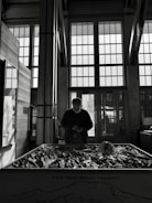 A person stands in front of a large model of Mount Fremont inside a building with large grid-patterned windows. The lighting casts a silhouette, emphasizing the interior architecture and the detailed topography of the model.