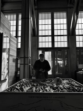 A person stands in front of a large model of Mount Fremont inside a building with large grid-patterned windows. The lighting casts a silhouette, emphasizing the interior architecture and the detailed topography of the model.