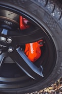 A close-up view of a car wheel featuring a black alloy rim with a visible red brake caliper labeled 'Brembo'. The tire tread is prominent and the background includes some foliage.
