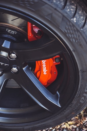 A close-up view of a car wheel featuring a black alloy rim with a visible red brake caliper labeled 'Brembo'. The tire tread is prominent and the background includes some foliage.