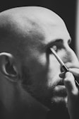 A monochrome image shows a bald person with facial hair applying makeup with a brush to their closed eye.