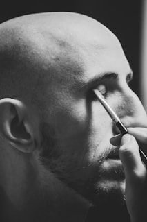 A monochrome image shows a bald person with facial hair applying makeup with a brush to their closed eye.
