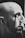 A monochrome image shows a bald person with facial hair applying makeup with a brush to their closed eye.
