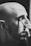 A monochrome image shows a bald person with facial hair applying makeup with a brush to their closed eye.