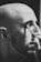 A monochrome image shows a bald person with facial hair applying makeup with a brush to their closed eye.