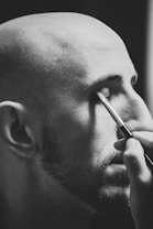 A monochrome image shows a bald person with facial hair applying makeup with a brush to their closed eye.