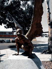 A bronze sculpture depicting a mythological creature with the body of a human and large wings. The figure is crouched on a stone pedestal in an outdoor setting surrounded by trees and parked cars. The scene includes strong shadows, suggesting it is a sunny day.