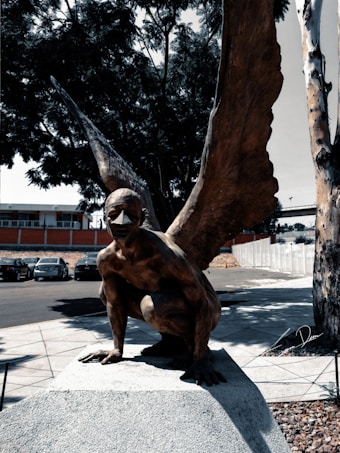 A bronze sculpture depicting a mythological creature with the body of a human and large wings. The figure is crouched on a stone pedestal in an outdoor setting surrounded by trees and parked cars. The scene includes strong shadows, suggesting it is a sunny day.