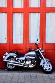 A silver motorcycle is parked in front of a red paneled door with glass windows. The bike is sleek and shiny, with chrome details and a black seat. The ground is made of light-colored paving stones.