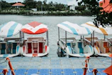 A row of colorful pedal boats is moored to a floating dock on a calm body of water. Each boat has a canopy, with distinct numbers and colors, including red, blue, and orange. The setting is serene, with lush greenery and a building visible in the background.