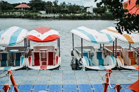 A row of colorful pedal boats is moored to a floating dock on a calm body of water. Each boat has a canopy, with distinct numbers and colors, including red, blue, and orange. The setting is serene, with lush greenery and a building visible in the background.