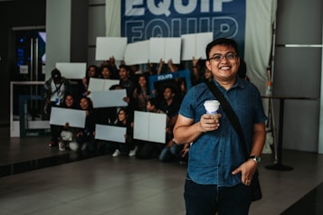 A group of people hold blank white boards and appear to be posing for a photo in a public indoor space. A smiling individual in the foreground holds a cup of coffee and stands slightly apart from the group. The atmosphere suggests an event or gathering, with a banner in the background displaying the word 'EQUIP'.