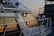 A close-up view of the side of a yacht showing its window reflecting the colors of a sunset. The metallic surface of the yacht gleams, and a part of the railing is visible. The background has another yacht slightly blurred, suggesting a marina setting at dusk.