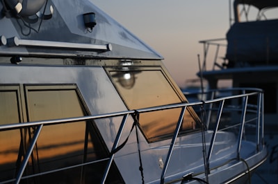 A close-up view of the side of a yacht showing its window reflecting the colors of a sunset. The metallic surface of the yacht gleams, and a part of the railing is visible. The background has another yacht slightly blurred, suggesting a marina setting at dusk.