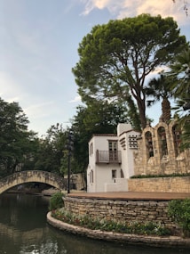 A picturesque scene featuring a stone bridge arching over a calm body of water, surrounded by lush greenery. To the right, a white building with a small balcony is situated next to an ornate stone wall with decorative arches and plant pots. A large, leafy tree extends over the scene, creating a serene and inviting atmosphere.