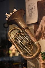 A brass instrument, likely a tuba, is mounted on a wall. The instrument has an elegant, polished surface with intricate tubing. Above it, a detailed sketch of a person's face is pinned to the wall, adding an artistic touch to the scene. The background is softly lit, creating a warm ambiance.
