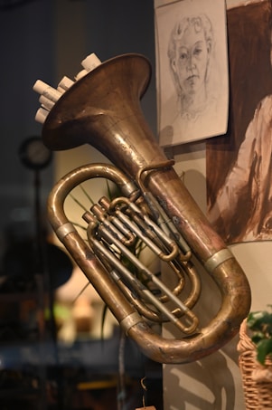 A brass instrument, likely a tuba, is mounted on a wall. The instrument has an elegant, polished surface with intricate tubing. Above it, a detailed sketch of a person's face is pinned to the wall, adding an artistic touch to the scene. The background is softly lit, creating a warm ambiance.