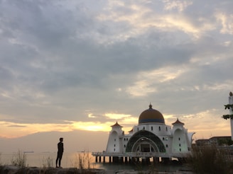 A mosque with a striking dome and intricate architectural design stands on the water's edge during sunset. The sky is partly cloudy, with hues of orange and yellow creating a serene atmosphere. A silhouette of a person stands in the foreground, surrounded by grass and rocks.
