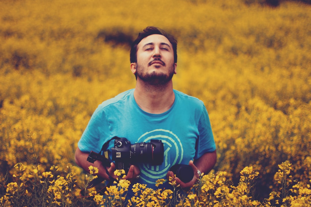man wearing gray crew-neck shirt holding black DSLR camera standing in the middle of yellow flowers