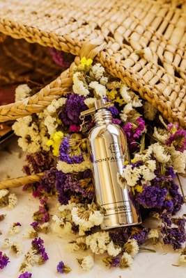 A silver bottle of Montale Paris Vanilla Absolu perfume is surrounded by a colorful array of dried flowers, including purple, yellow, and white blossoms. The scene includes a woven wicker or straw mat positioned behind the bottle and flowers, adding a rustic and natural feel.