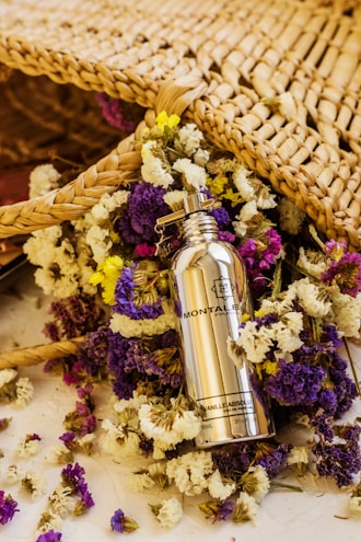 A silver bottle of Montale Paris Vanilla Absolu perfume is surrounded by a colorful array of dried flowers, including purple, yellow, and white blossoms. The scene includes a woven wicker or straw mat positioned behind the bottle and flowers, adding a rustic and natural feel.