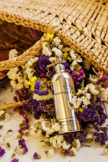 A silver bottle of Montale Paris Vanilla Absolu perfume is surrounded by a colorful array of dried flowers, including purple, yellow, and white blossoms. The scene includes a woven wicker or straw mat positioned behind the bottle and flowers, adding a rustic and natural feel.