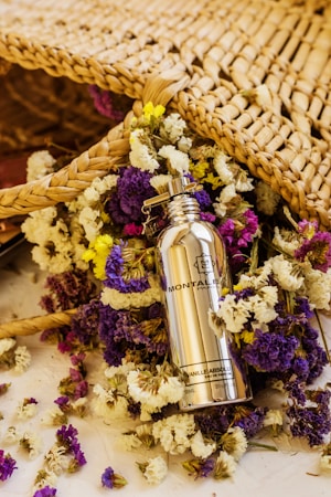 A silver bottle of Montale Paris Vanilla Absolu perfume is surrounded by a colorful array of dried flowers, including purple, yellow, and white blossoms. The scene includes a woven wicker or straw mat positioned behind the bottle and flowers, adding a rustic and natural feel.