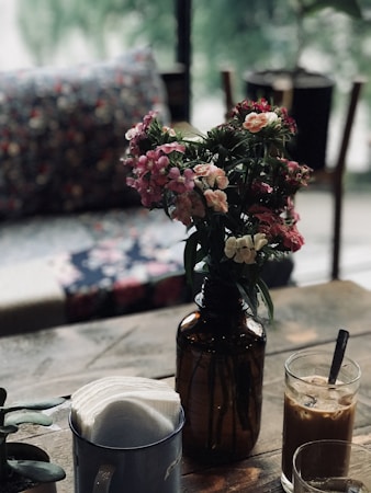 A cozy setting with a vase of pink and white flowers on a wooden table, next to a glass of iced coffee and a cup containing napkins. The background features a blurred view of potted plants and a couch with a colorful cushion, creating a relaxed atmosphere.