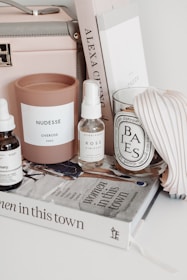 A neatly arranged collection of items on a white surface, including a pink candle labeled 'NUDESSE' by Overose Paris, a spray bottle labeled 'ROSE' by Herbivore, a scented candle in a glass labeled 'BAIES' by diptyque, a tan box with a handle, and a stack of books. The titles of the books include 'Women in This Town' and another spine with 'ALEXA CHUNG.' There is also a pleated item next to the books.