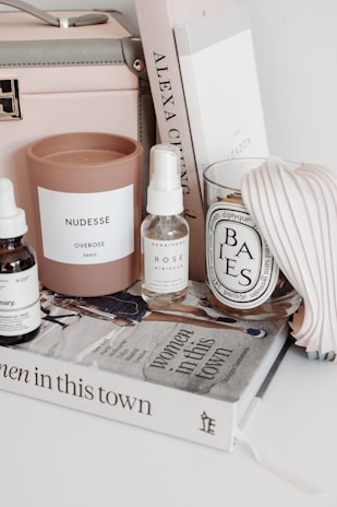 A neatly arranged collection of items on a white surface, including a pink candle labeled 'NUDESSE' by Overose Paris, a spray bottle labeled 'ROSE' by Herbivore, a scented candle in a glass labeled 'BAIES' by diptyque, a tan box with a handle, and a stack of books. The titles of the books include 'Women in This Town' and another spine with 'ALEXA CHUNG.' There is also a pleated item next to the books.