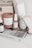 A neatly arranged collection of items on a white surface, including a pink candle labeled 'NUDESSE' by Overose Paris, a spray bottle labeled 'ROSE' by Herbivore, a scented candle in a glass labeled 'BAIES' by diptyque, a tan box with a handle, and a stack of books. The titles of the books include 'Women in This Town' and another spine with 'ALEXA CHUNG.' There is also a pleated item next to the books.