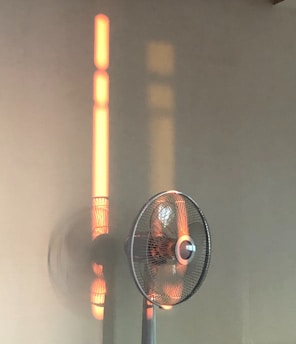 A fan stands against a wall with soft orange light creating a shadow. The light forms distinct vertical bands, casting an interesting pattern that coincides with the rotating blades. The overall ambiance feels calm and serene.
