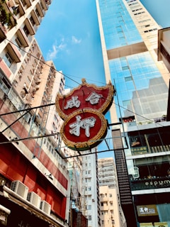 A vibrant urban setting with tall buildings surrounding a prominent red and gold sign featuring Chinese characters. The sign hangs amidst high-rise structures, with modern glass facades and more traditional apartment-style buildings visible. The sky is clear and blue, adding contrast to the scene.