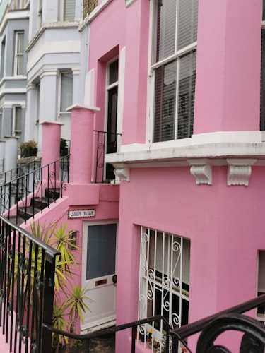 A row of vibrant, pastel-colored houses with a prominent pink facade. The building features a black wrought iron railing leading to a white door. Large windows with white trim and decorative moldings overlook the street. A small staircase ascends beside the building, lined by plants.