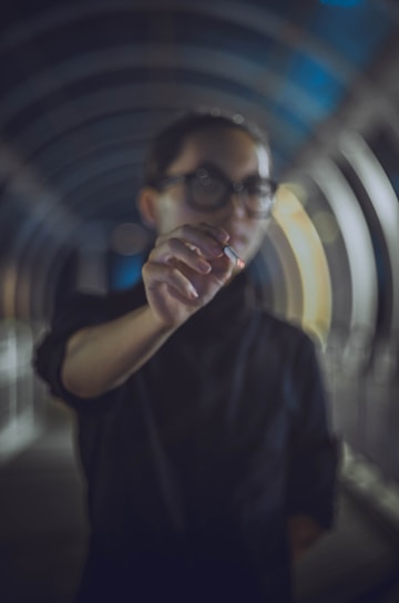 A person with glasses is holding a lit cigarette while standing in a dimly lit, arched tunnel. The background features blurred lines of the tunnel structure, creating a sense of depth and focus on the cigarette.