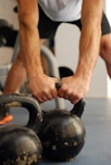 A person is gripping two kettlebells placed on a gym floor. The focus is on the person's forearms and hands, with the kettlebells showing signs of wear. The background is blurred, indicating a gym setting.
